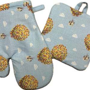 May include: A light blue oven mitt and pot holder set. The set features a pattern of colorful balloon clusters, white clouds, and small houses. The mitt has a loop for hanging. The pot holder has a loop for hanging.