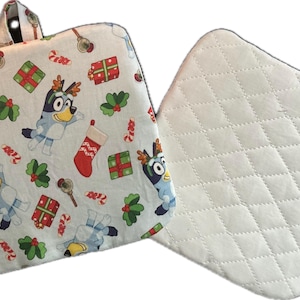 May include: A Christmas-themed pot holder with a white background featuring cartoon characters, red stockings, green holly, and wrapped gifts. The reverse side is white with a quilted pattern. Includes a fabric loop for hanging.