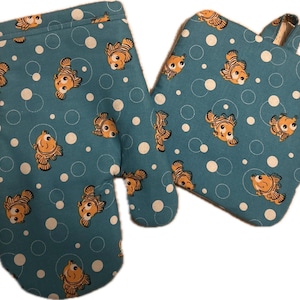 May include: A pair of teal oven mitts and pot holder with a repeating pattern of orange cartoon fish and white polka dots. The mitt has a loop for hanging. The pot holder is square.