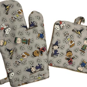 May include: A set of two grey oven mitts with a Halloween theme. The mitts feature a repeating pattern of Peanuts characters dressed in costumes and jack-o-lanterns.