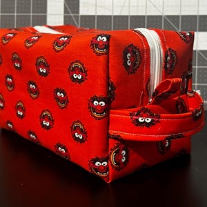 May include: A red, rectangular cosmetic bag featuring a repeating pattern of a cartoon character's face. The bag has a white zipper, a matching handle, and a white zipper pull. The bag is sitting on a black surface.