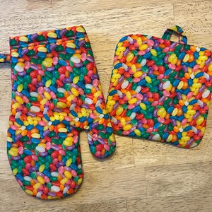 May include: A colorful oven mitt and pot holder set. The fabric is covered in a pattern of jelly beans in various colors, including red, yellow, green, purple, and orange. The oven mitt has a loop for hanging.