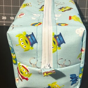 Peut inclure: Une pochette rectangulaire en tissu bleu clair avec une fermeture &eacute;clair blanche. La pochette est orn&eacute;e d'un motif r&eacute;p&eacute;t&eacute; d'aliens verts dans diverses poses. La fermeture &eacute;clair est au centre.