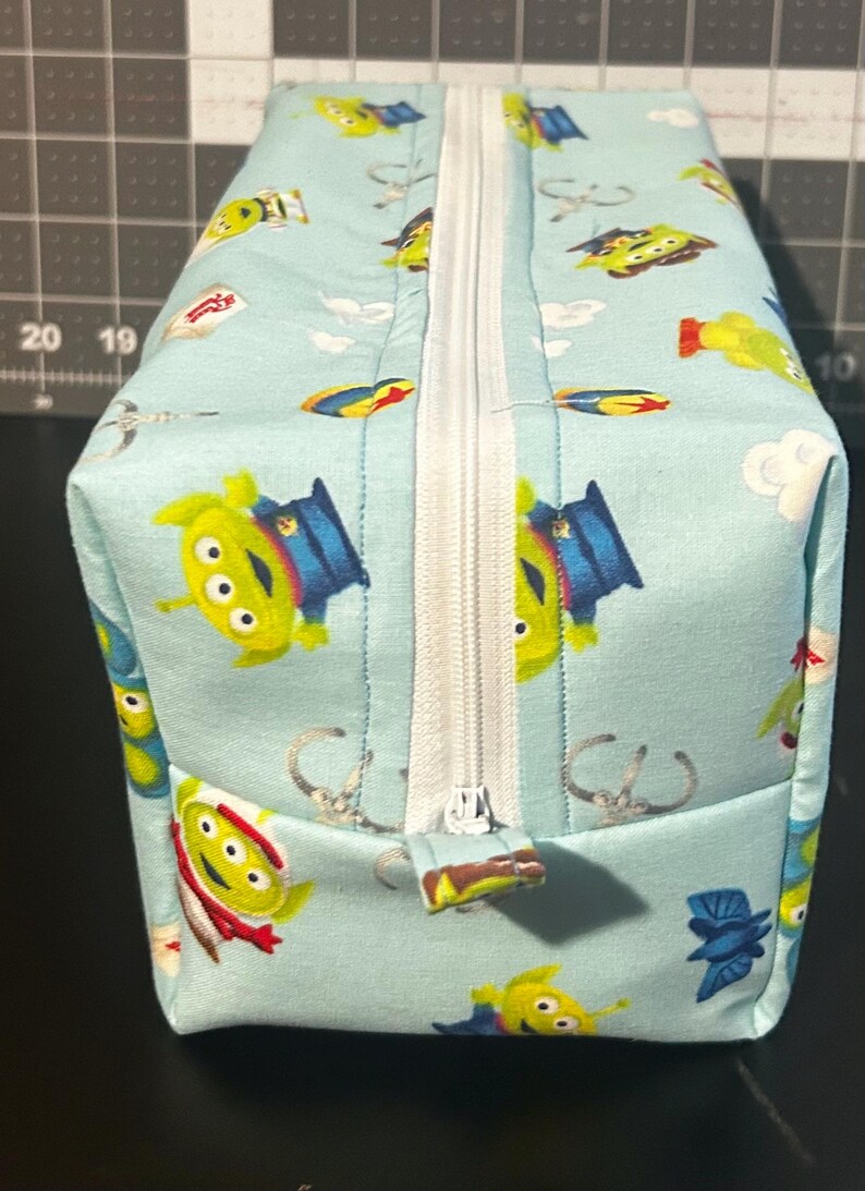 Peut inclure: Une pochette rectangulaire en tissu bleu clair avec une fermeture &eacute;clair blanche. La pochette est orn&eacute;e d'un motif r&eacute;p&eacute;t&eacute; d'aliens verts dans diverses poses. La fermeture &eacute;clair est au centre.