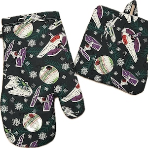 May include: A dark gray oven mitt and pot holder set with a repeating pattern of spaceships, Death Star ornaments, snowflakes, and pine needles. The spaceships are white, red, and purple. The oven mitt has a loop for hanging.
