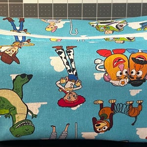 Puede incluir: Un neceser rectangular azul claro con un estampado repetido de personajes de Toy Story. La bolsa tiene un cierre de cremallera blanco y un asa peque&ntilde;a en un lado. Los personajes incluyen a Woody, Buzz Lightyear y Mr. Potato Head.