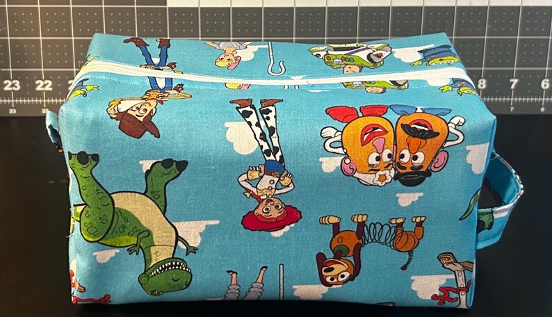 Puede incluir: Un neceser rectangular azul claro con un estampado repetido de personajes de Toy Story. La bolsa tiene un cierre de cremallera blanco y un asa peque&ntilde;a en un lado. Los personajes incluyen a Woody, Buzz Lightyear y Mr. Potato Head.