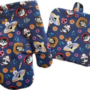 May include: A blue oven mitt and a matching pot holder with a colorful pattern of characters from the movie Coco. The characters are wearing traditional Mexican clothing and have sugar skulls on their faces.