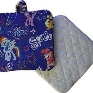 May include: A blue pot holder with a loop, featuring cartoon ponies, stars, and the words "Love" and "Star" in white. A separate white quilted pad is next to the pot holder. The pot holder has a colorful design with a rainbow and a hot air balloon.