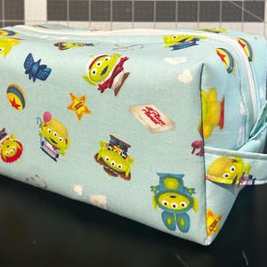 Peut inclure: Une trousse de toilette rectangulaire bleu clair avec un motif r&eacute;p&eacute;t&eacute; d'extraterrestres de dessins anim&eacute;s, d'&eacute;toiles et d'autres graphiques de personnages. Le sac a une fermeture &eacute;clair blanche et une petite poign&eacute;e en tissu. Le th&egrave;me g&eacute;n&eacute;ral est un film d'animation pour enfants.