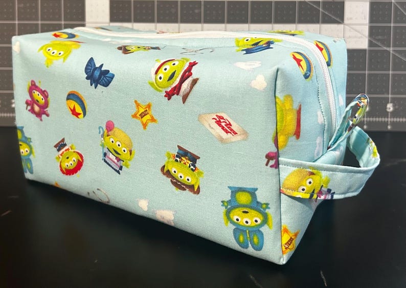 Peut inclure: Une trousse de toilette rectangulaire bleu clair avec un motif r&eacute;p&eacute;t&eacute; d'extraterrestres de dessins anim&eacute;s, d'&eacute;toiles et d'autres graphiques de personnages. Le sac a une fermeture &eacute;clair blanche et une petite poign&eacute;e en tissu. Le th&egrave;me g&eacute;n&eacute;ral est un film d'animation pour enfants.