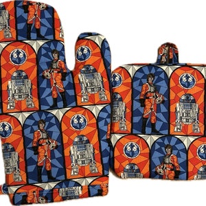 May include: A set of two Star Wars themed oven mitts. The mitts feature a stained glass design with images of R2-D2 and a character in a flight suit. The mitts are orange, blue, and white.