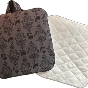 May include: Two quilted pot holders. One is dark gray with a repeating pattern of ornate designs and the letters "HM". The other is white with a diamond pattern. One has a loop for hanging.