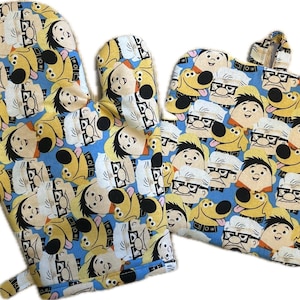 May include: A blue oven mitt and pot holder set. The set features a repeating pattern of cartoon characters in yellow, white, and brown. The oven mitt is on the left, and the pot holder is on the right with a hanging loop.