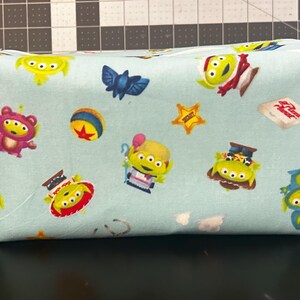 Peut inclure: Une trousse de toilette rectangulaire en tissu bleu clair avec un motif r&eacute;p&eacute;t&eacute; d'extraterrestres de dessins anim&eacute;s, d'une chauve-souris bleue, d'une balle et d'une &eacute;toile. Le sac a une fermeture &eacute;clair et de petites boucles en tissu &agrave; chaque extr&eacute;mit&eacute;.