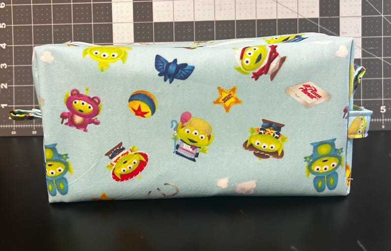 Peut inclure: Une trousse de toilette rectangulaire en tissu bleu clair avec un motif r&eacute;p&eacute;t&eacute; d'extraterrestres de dessins anim&eacute;s, d'une chauve-souris bleue, d'une balle et d'une &eacute;toile. Le sac a une fermeture &eacute;clair et de petites boucles en tissu &agrave; chaque extr&eacute;mit&eacute;.