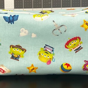 Peut inclure: Une trousse de maquillage rectangulaire en tissu bleu clair avec un motif r&eacute;p&eacute;t&eacute; de personnages de dessins anim&eacute;s, dont des extraterrestres, des &eacute;toiles et une balle. Le sac a une fermeture &eacute;clair et de petites boucles en tissu sur les c&ocirc;t&eacute;s.