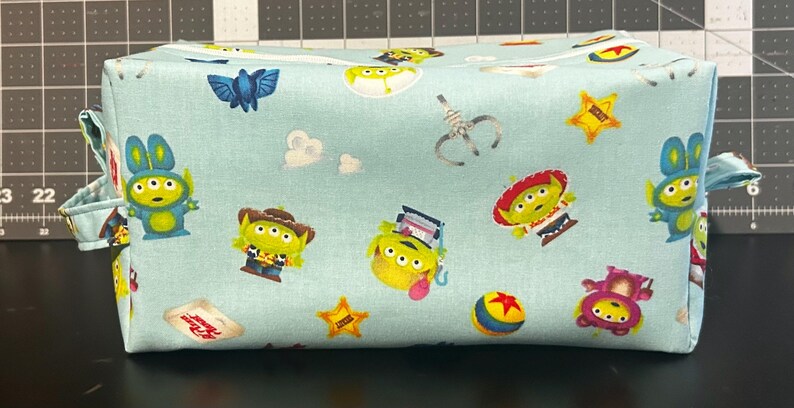 Peut inclure: Une trousse de maquillage rectangulaire en tissu bleu clair avec un motif r&eacute;p&eacute;t&eacute; de personnages de dessins anim&eacute;s, dont des extraterrestres, des &eacute;toiles et une balle. Le sac a une fermeture &eacute;clair et de petites boucles en tissu sur les c&ocirc;t&eacute;s.