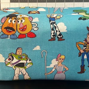 Puede incluir: Una bolsa rectangular de tela azul claro con personajes de dibujos animados de la franquicia Toy Story. La bolsa tiene un asa en un lado y est&aacute; decorada con im&aacute;genes de Woody, Buzz Lightyear y otros personajes.