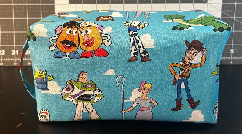 Puede incluir: Una bolsa rectangular de tela azul claro con personajes de dibujos animados de la franquicia Toy Story. La bolsa tiene un asa en un lado y est&aacute; decorada con im&aacute;genes de Woody, Buzz Lightyear y otros personajes.