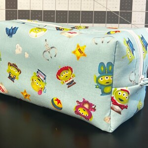 Peut inclure: Une pochette rectangulaire en tissu bleu clair avec une fermeture &eacute;clair blanche. La pochette est recouverte d'un motif r&eacute;p&eacute;t&eacute; d'aliens de dessins anim&eacute;s, d'&eacute;toiles et d'autres graphiques th&eacute;matiques. La pochette est pos&eacute;e sur une surface noire.