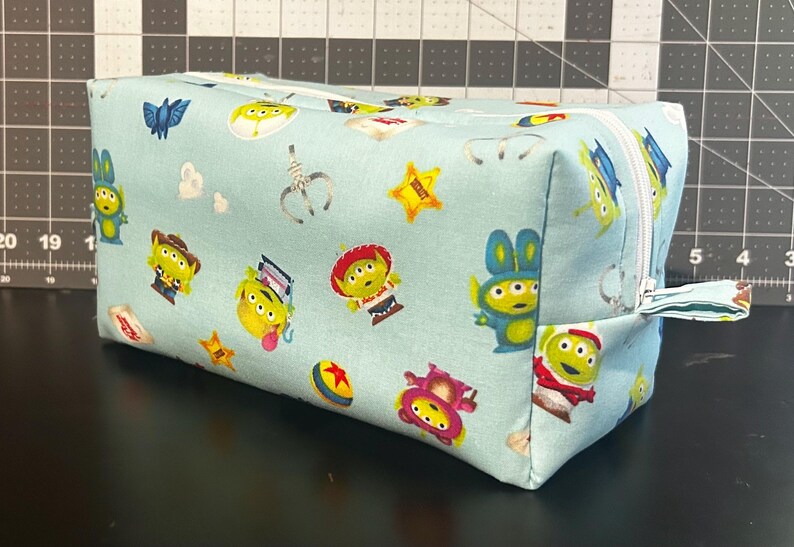 Peut inclure: Une pochette rectangulaire en tissu bleu clair avec une fermeture &eacute;clair blanche. La pochette est recouverte d'un motif r&eacute;p&eacute;t&eacute; d'aliens de dessins anim&eacute;s, d'&eacute;toiles et d'autres graphiques th&eacute;matiques. La pochette est pos&eacute;e sur une surface noire.