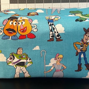 Puede incluir: Una bolsa rectangular de tela azul claro con personajes de dibujos animados de la franquicia Toy Story. La bolsa tiene cierre de cremallera y un asa peque&ntilde;a. Los personajes incluyen a Mr. Potato Head, Buzz Lightyear y Woody.