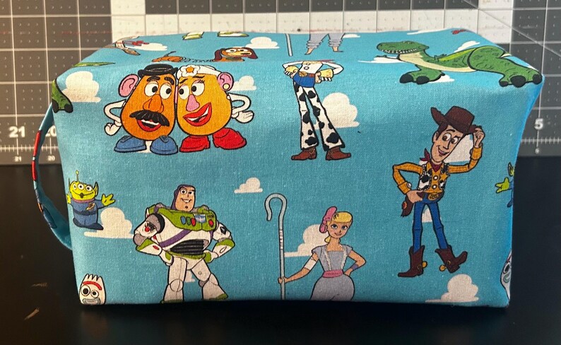 Puede incluir: Una bolsa rectangular de tela azul claro con personajes de dibujos animados de la franquicia Toy Story. La bolsa tiene cierre de cremallera y un asa peque&ntilde;a. Los personajes incluyen a Mr. Potato Head, Buzz Lightyear y Woody.