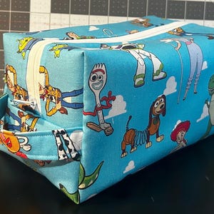 Puede incluir: Una bolsa rectangular con cremallera con un fondo azul claro y un patr&oacute;n repetido de personajes de dibujos animados. Los personajes incluyen un dinosaurio verde, un perro muelle y un tenedor. La bolsa tiene una cremallera blanca y un asa peque&ntilde;a.