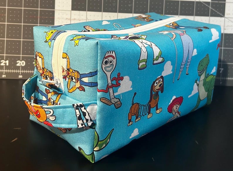 Puede incluir: Una bolsa rectangular con cremallera con un fondo azul claro y un patr&oacute;n repetido de personajes de dibujos animados. Los personajes incluyen un dinosaurio verde, un perro muelle y un tenedor. La bolsa tiene una cremallera blanca y un asa peque&ntilde;a.