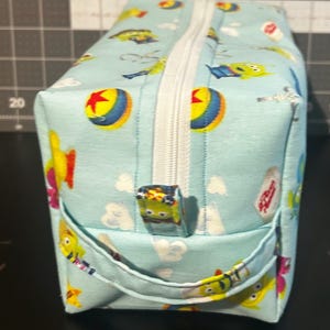 Peut inclure: Une trousse de maquillage rectangulaire bleu clair avec une fermeture &eacute;clair blanche et une petite poign&eacute;e. Le sac est d&eacute;cor&eacute; de personnages de dessins anim&eacute;s, de nuages et d'un motif de balle jaune et rouge.