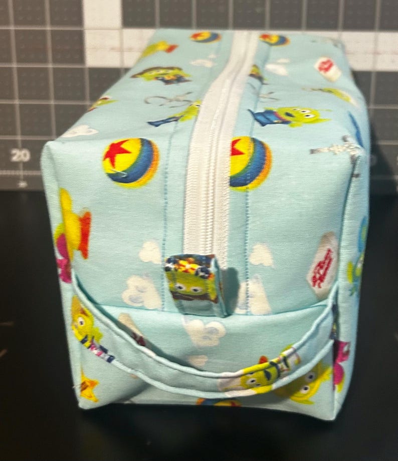 Peut inclure: Une trousse de maquillage rectangulaire bleu clair avec une fermeture &eacute;clair blanche et une petite poign&eacute;e. Le sac est d&eacute;cor&eacute; de personnages de dessins anim&eacute;s, de nuages et d'un motif de balle jaune et rouge.