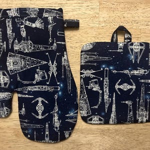 May include: Navy blue oven mitt and pot holder set. The set features a white schematic design of various space ships and vehicles against a starry background. The oven mitt has a loop for hanging.