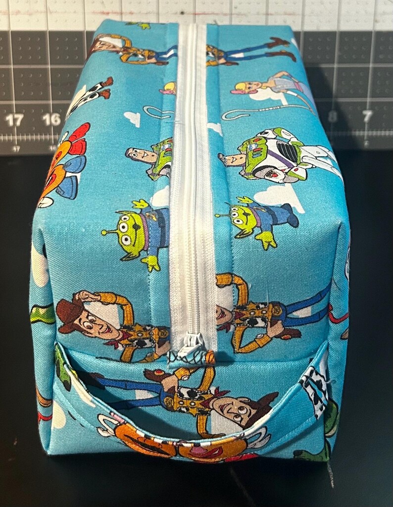 Puede incluir: Un neceser rectangular azul claro con cremallera blanca y un peque&ntilde;o bolsillo. La bolsa est&aacute; decorada con un patr&oacute;n repetido de personajes de dibujos animados, incluyendo Woody, Buzz Lightyear y los Little Green Men. La bolsa est&aacute; hecha de una tela resistente.