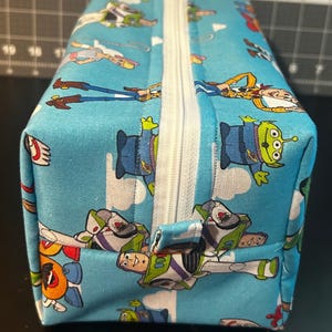 Puede incluir: Una bolsa rectangular con cremallera con un fondo azul claro y un patr&oacute;n repetido de personajes de dibujos animados. Los personajes incluyen un vaquero, un guardabosques espacial y un extraterrestre de tres ojos. La cremallera es blanca.