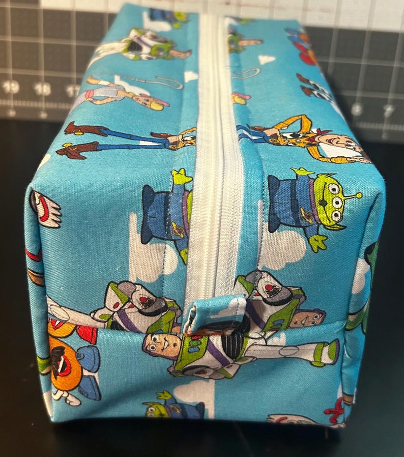 Puede incluir: Una bolsa rectangular con cremallera con un fondo azul claro y un patr&oacute;n repetido de personajes de dibujos animados. Los personajes incluyen un vaquero, un guardabosques espacial y un extraterrestre de tres ojos. La cremallera es blanca.