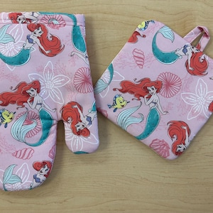 May include: A pink oven mitt and pot holder set featuring a mermaid design. The mermaid has red hair and a teal tail. The set is decorated with flowers, shells, and a yellow fish.