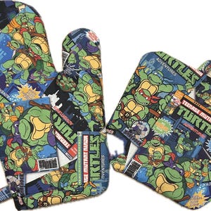 May include: A set of two Teenage Mutant Ninja Turtles oven mitts. The mitts are made of fabric and feature a comic book design with the Teenage Mutant Ninja Turtles characters. The mitts are blue, green, red, yellow, and black.