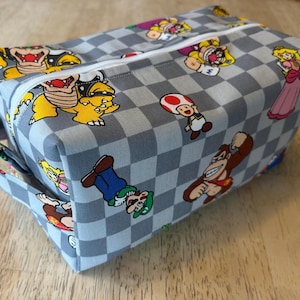 May include: A rectangular, gray and white checkered fabric bag featuring colorful cartoon characters. The bag has a white zipper and a small handle. Characters include Bowser, Princess Peach, and Donkey Kong.