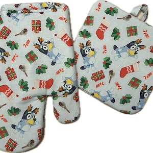 May include: A light blue oven mitt and pot holder set with a festive pattern. The pattern includes cartoon characters, wrapped gifts, holly, candy canes, and Christmas stockings. The oven mitt has a loop for hanging.