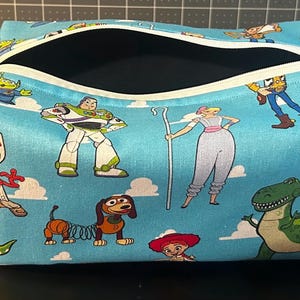 Puede incluir: Un neceser rectangular con cierre de cremallera. La bolsa presenta un fondo azul claro con personajes de dibujos animados de la pel&iacute;cula Toy Story. La bolsa tiene un asa peque&ntilde;a en el lateral.