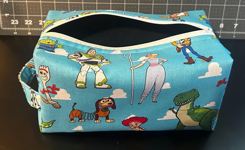 Puede incluir: Un neceser rectangular con cierre de cremallera. La bolsa presenta un fondo azul claro con personajes de dibujos animados de la pel&iacute;cula Toy Story. La bolsa tiene un asa peque&ntilde;a en el lateral.