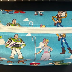 Puede incluir: Una bolsa rectangular con cremallera y fondo azul claro con personajes de dibujos animados. La bolsa tiene una cremallera blanca y un asa peque&ntilde;a en un lado. Los personajes incluyen a Woody, Buzz Lightyear y otros personajes de Toy Story.