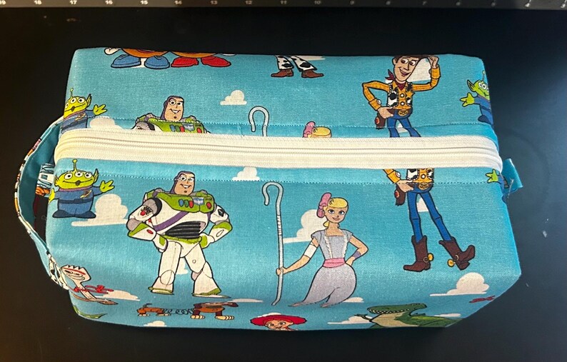 Puede incluir: Una bolsa rectangular con cremallera y fondo azul claro con personajes de dibujos animados. La bolsa tiene una cremallera blanca y un asa peque&ntilde;a en un lado. Los personajes incluyen a Woody, Buzz Lightyear y otros personajes de Toy Story.