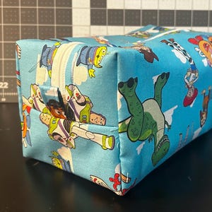 Puede incluir: Una bolsa rectangular con cremallera con un fondo azul claro y un patr&oacute;n repetido de personajes de Toy Story. La bolsa tiene una cremallera blanca y est&aacute; hecha de tela. Los personajes incluyen a Buzz Lightyear, Woody y Rex.