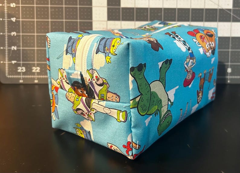 Puede incluir: Una bolsa rectangular con cremallera con un fondo azul claro y un patr&oacute;n repetido de personajes de Toy Story. La bolsa tiene una cremallera blanca y est&aacute; hecha de tela. Los personajes incluyen a Buzz Lightyear, Woody y Rex.