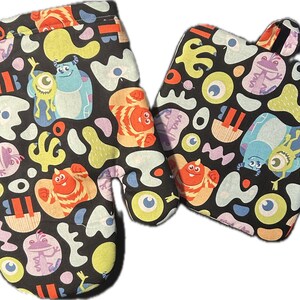 May include: A set of two black oven mitts with a colorful pattern featuring characters from the movie Monsters Inc. The pattern includes green, purple, orange, yellow, and blue characters on a black background.