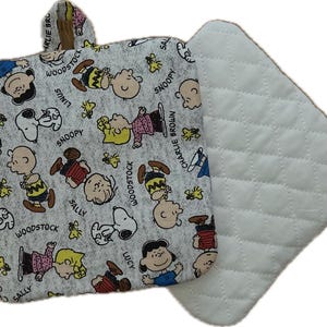 May include: A gray and white Peanuts-themed potholder and matching square pad. The potholder features a loop for hanging and a pattern of Snoopy, Charlie Brown, Woodstock, and other characters. The pad is white with a quilted design.