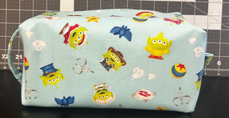 Peut inclure: Une trousse de maquillage rectangulaire bleu clair avec un motif r&eacute;p&eacute;t&eacute; de personnages de Toy Story. Le sac comprend des images d'aliens, de Woody, de Buzz Lightyear et d'autres personnages. Il a une petite poign&eacute;e sur le c&ocirc;t&eacute;.