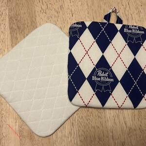 May include: Two quilted pot holders. One is solid white with a diamond pattern. The other has a blue and white argyle pattern with red stitching and the "Pabst Blue Ribbon" logo. One pot holder has a hanging loop.